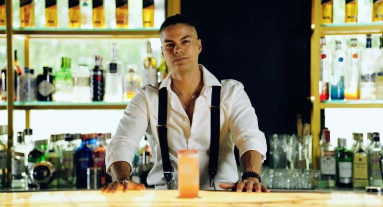 Barman en chemise blanche derriere un comptoir garni de bouteilles avec un cocktail servi