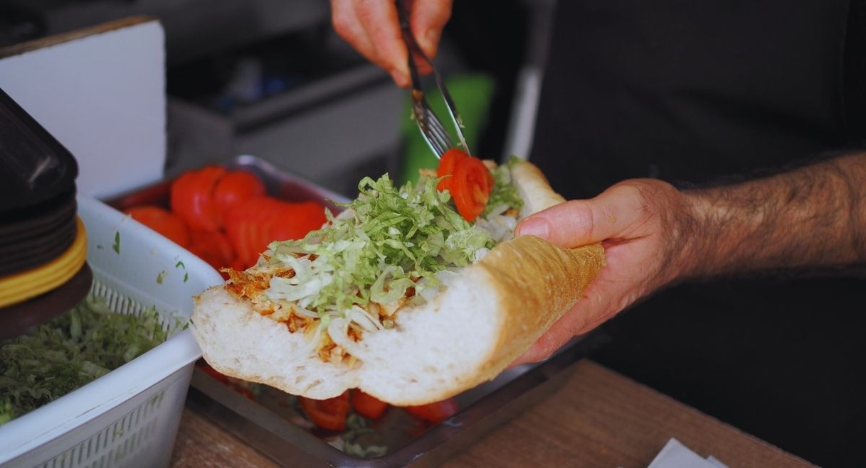 Mains garnissant un sandwich baguette de salade et de tomates a un comptoir de restauration